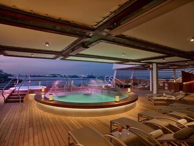 Outdoor whirlpools and with purple and pink lighting, water fountains cascading into the pools, deck chairs lined around them on deck with ocean in background.