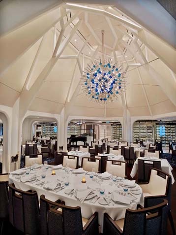 Seabourn Encore Dining room - Formal dining room, walls draped in white silk, with a large modern crystal chandelere above oval tables, set in fine fashion with linen napkins folded gracefully adorning fine china and silverware.