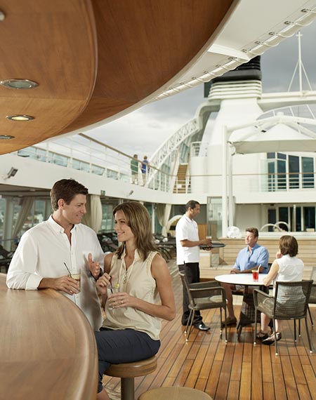 Man and woman smiling and enjoying cocktails at the Patio Bar. Another couple being served by crew at a table in the background.